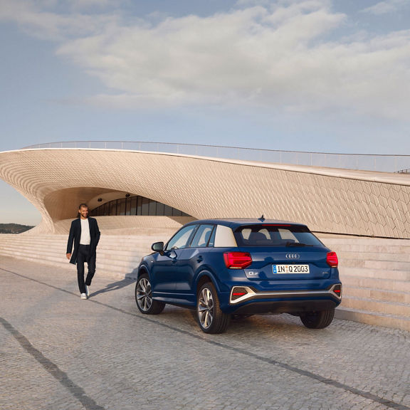 A blue Audi Q2 parked beside a modern wavy architecture with a walking person to the side.