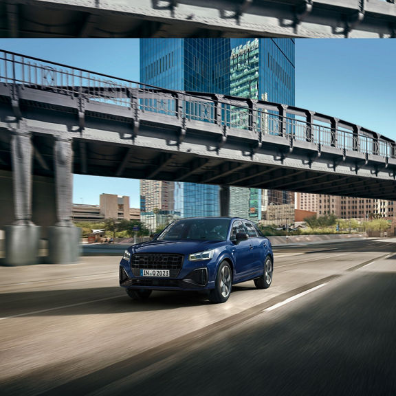 Blue SUV driving on a sunny city street under an overpass.