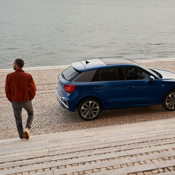 A person walking by a blue SUV parked near a water body, on a paved promenade.