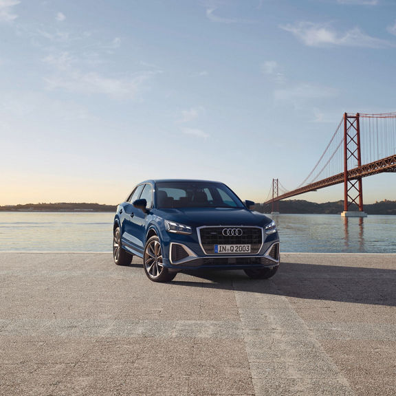 Blue car parked in front of a suspension bridge over water at sunset.