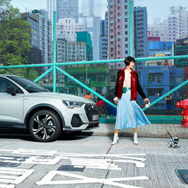 Full outdoor shot of a stylish woman standing in front of a light grey Audi SUV and a wire mesh fence that separates a basketball court from a city street.