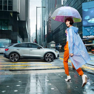 Full outdoor shot a person in orange pants and blue raincoat walks beside a light grey Audi Q3 on a city street.