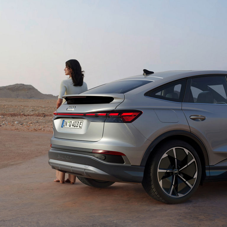 Rear and side view of Audi A4 in desert space with a lady stood behind gazing into mountain landscape.