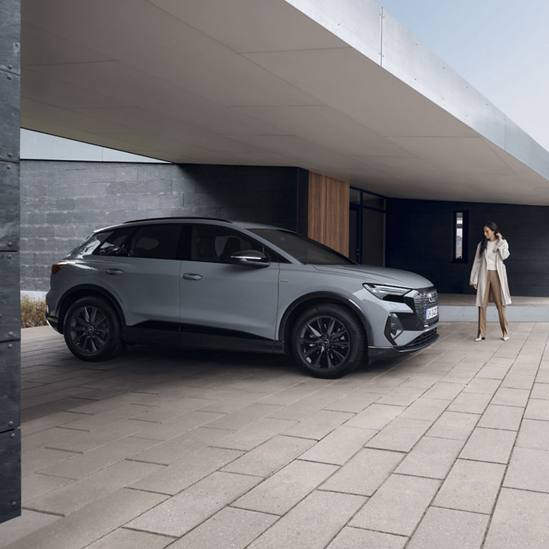 Lady walking infront of grey Audi Q4 e-tron with city buildings in the background.