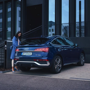 A blue Audi plugged in and charging outside a modern building.