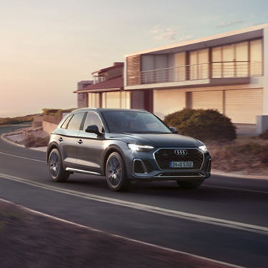 Audi SUV driving on a coastal road near modern architecture at sunset.