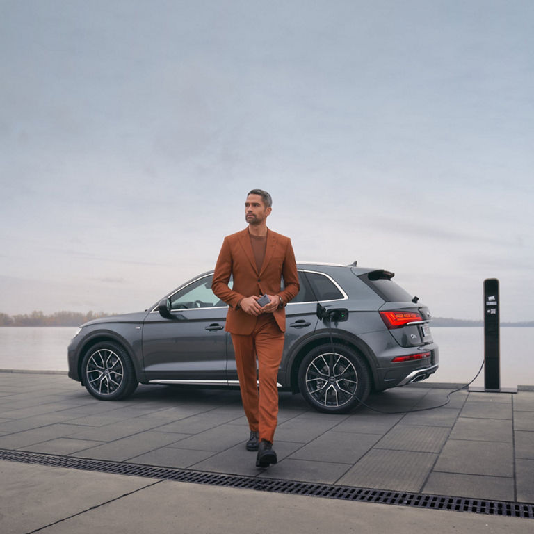 Person in a suit standing next to an electric SUV being charged at a waterfront station.