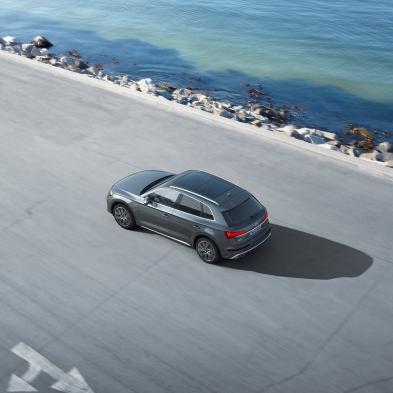 Aerial view of a grey SUV driving on a coastal road next to a calm sea.