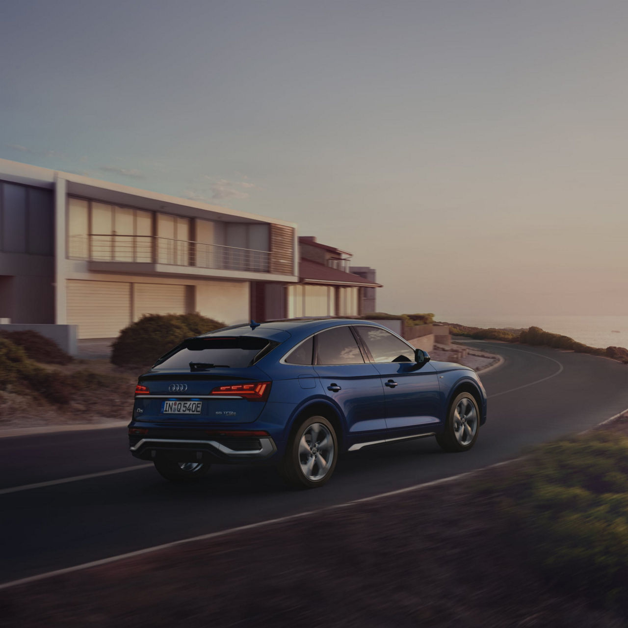 Blue car driving along a coastal road at dusk with modern houses in the background.