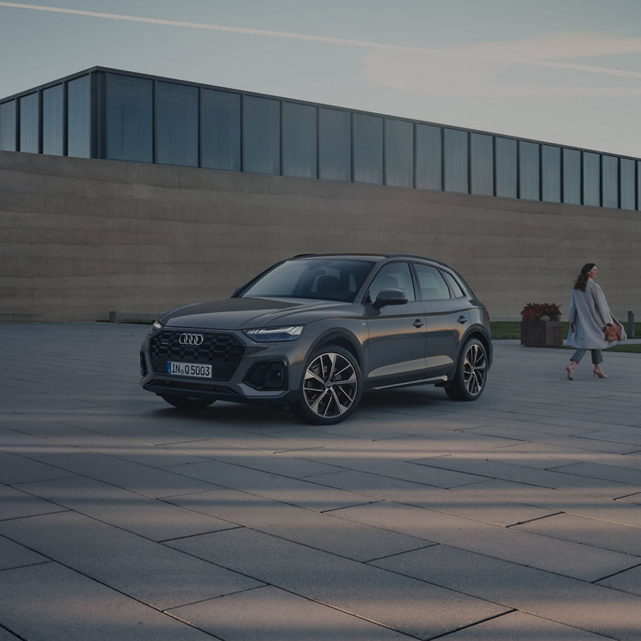 A grey Audi SUV parked on a plaza with a person walking in the background.