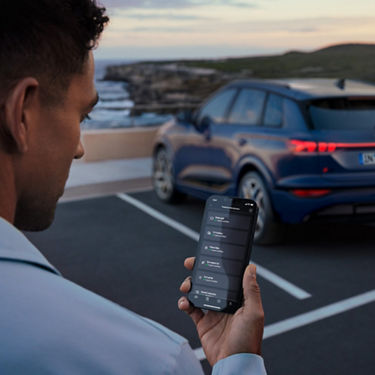 Person holding a smartphone with an app open in front of a parked blue SUV near the coast at dusk.