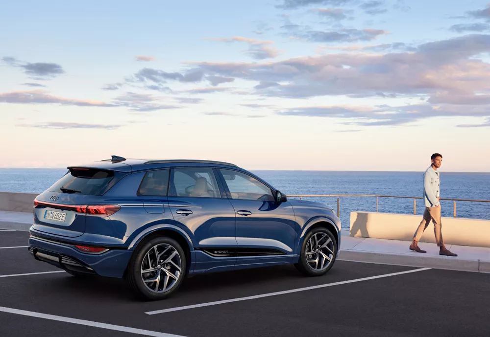  A blue Audi vehicle parked in a coastal setting, sea in the distance & a man walking to the right