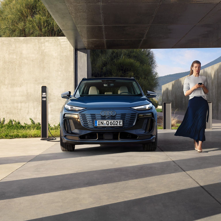 A woman walks past a parked Audi with a charger, focused on her phone.