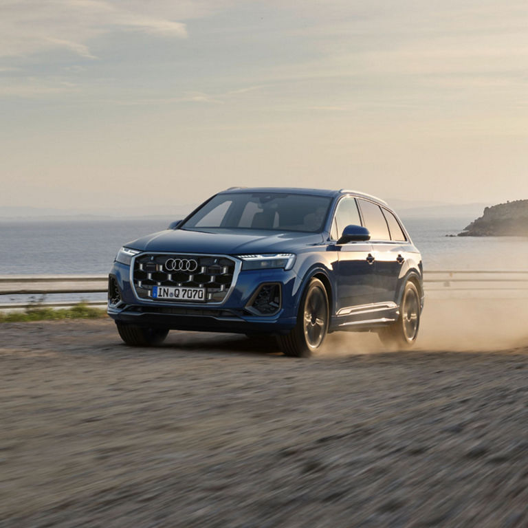 A blue SUV driving on a coastal road raising dust.