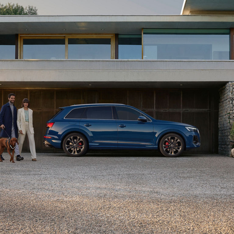 Blue SUV parked outside a modern house with a couple and a dog walking by.