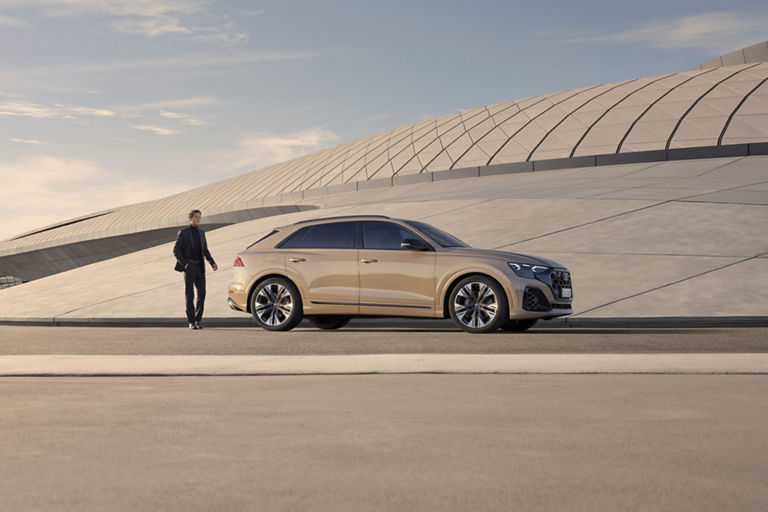 Man standing behind beige coloured Audi Q8 SUV with modern architecture in background.