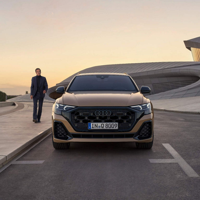 A man in a suit walking alongside a parked Audi S8 SUV, with modern architecture in the background.