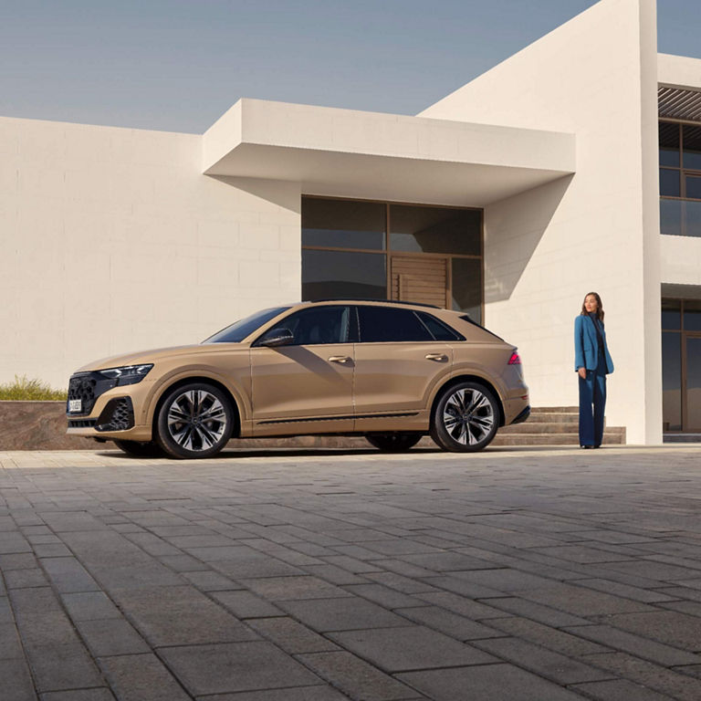 A luxury beige Audi SUV parked beside a modern building with a woman standing nearby.