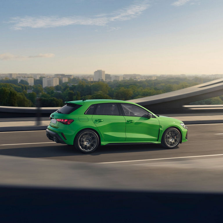 Bright green Audi RS3 Sportback driving on highway with city skyline in the background.