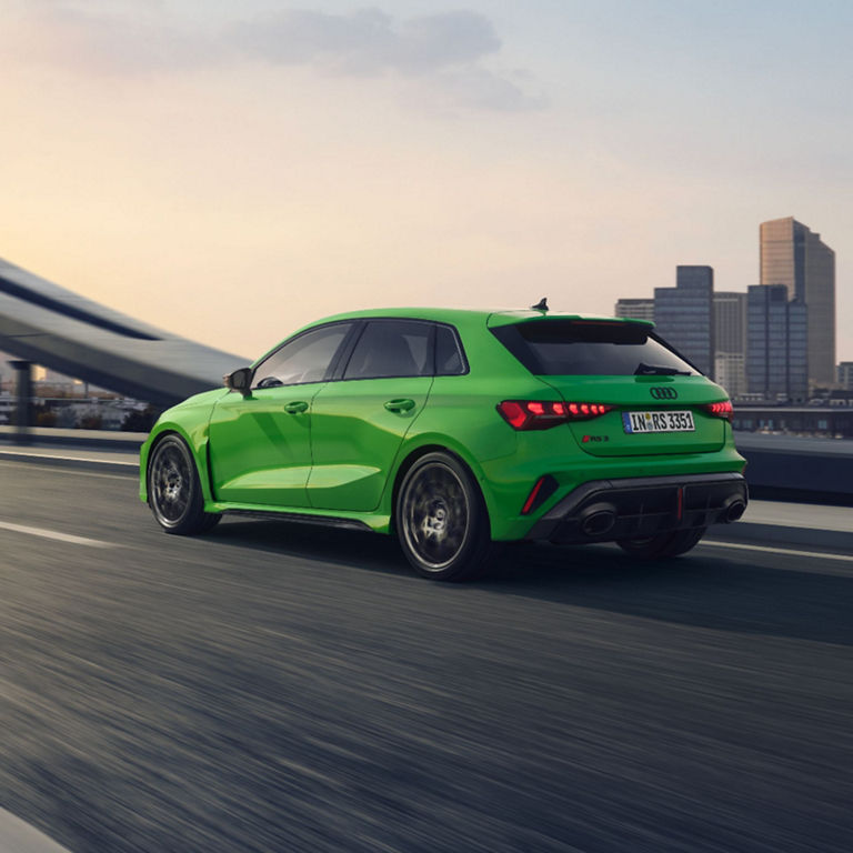 Green Audi RS3 Sportback driving on the highway with city skyline in the background.