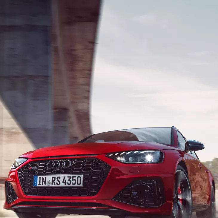 Red Audi car driving under a bridge, focus on the front grille and headlight.