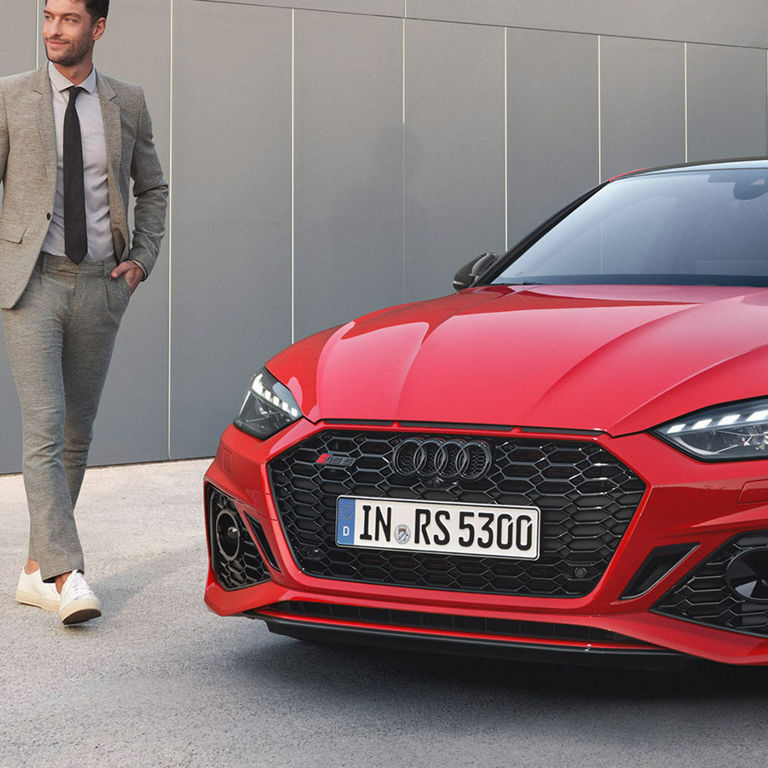 A person in business attire standing beside a red Audi car.