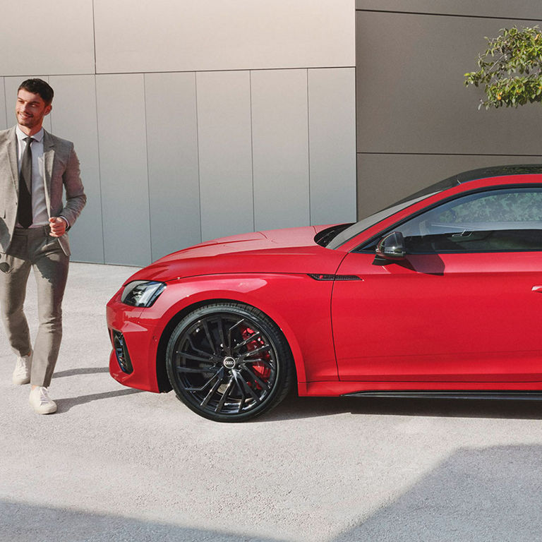 A person in a suit standing next to a red Audi car with black rims.