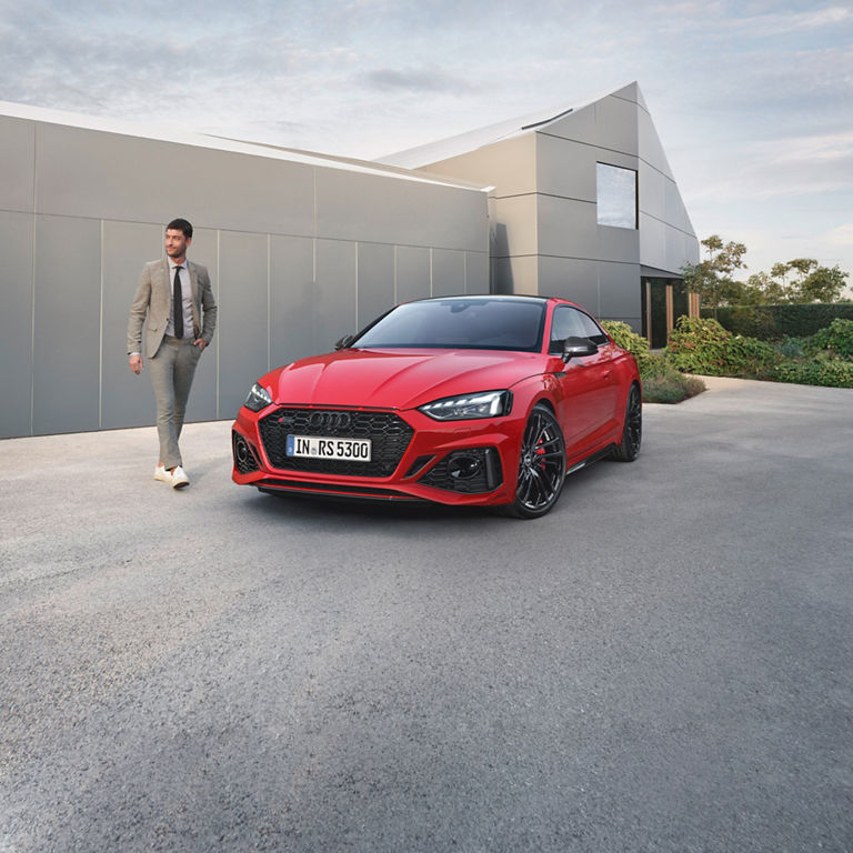 A red Audi RS5 Coupe parked outside a modern building with a person approaching it.