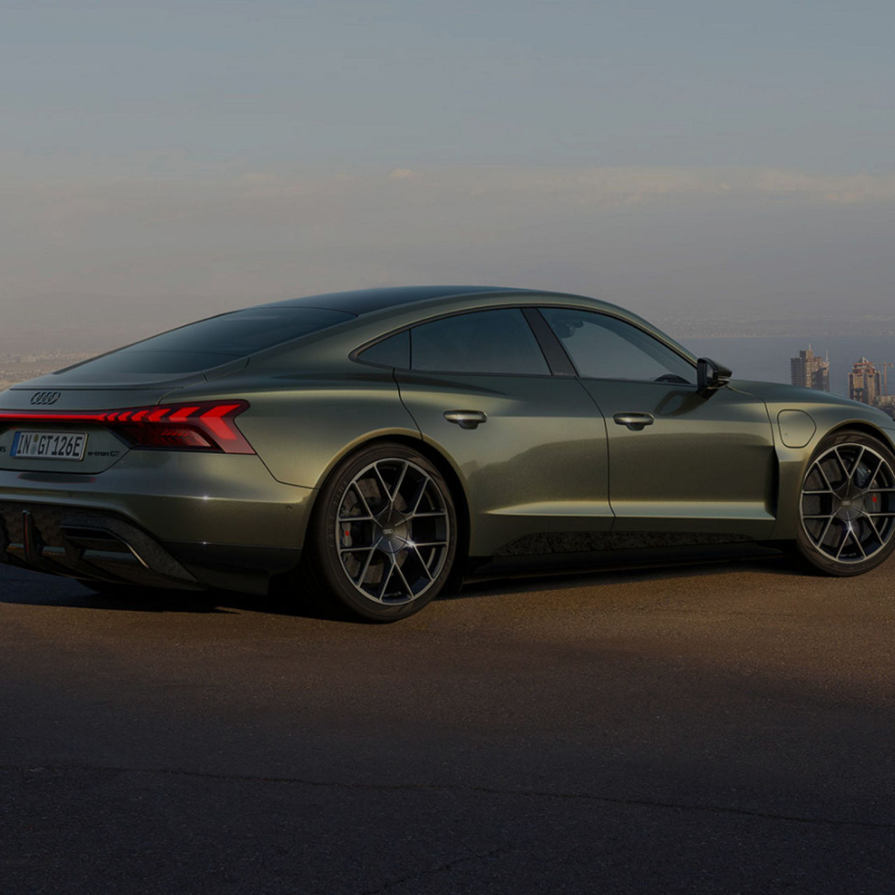 Side view of a olive green Audi RS e-tron GT performance against a blurred cityscape backdrop.