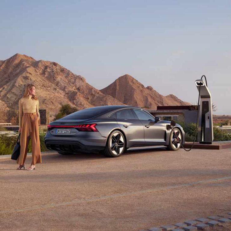 A person stands next to an electric car charging at a station with mountains in the background.