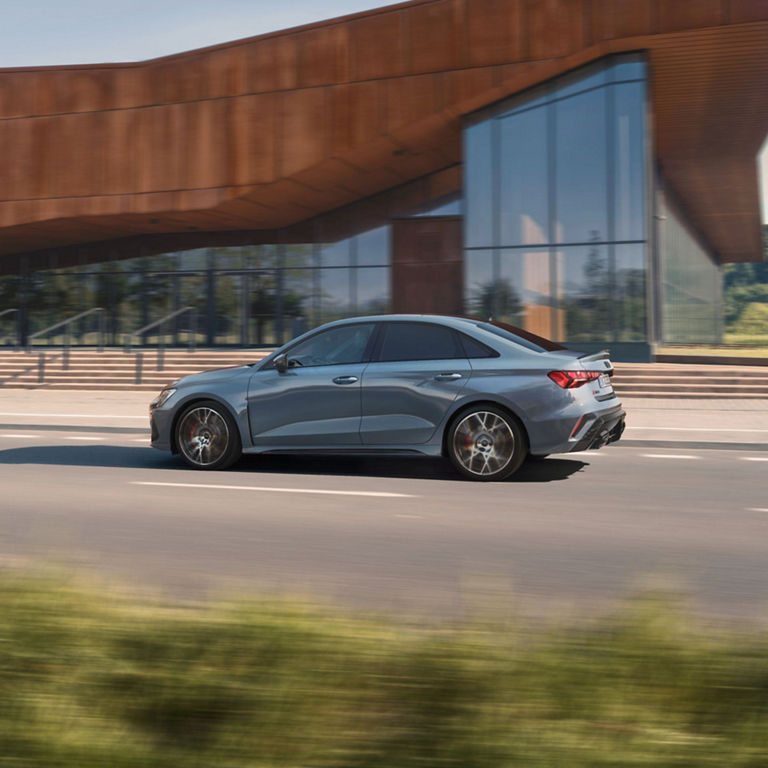 A silver Audi RS3 Saloon car in motion on a road with a modern building in the background.