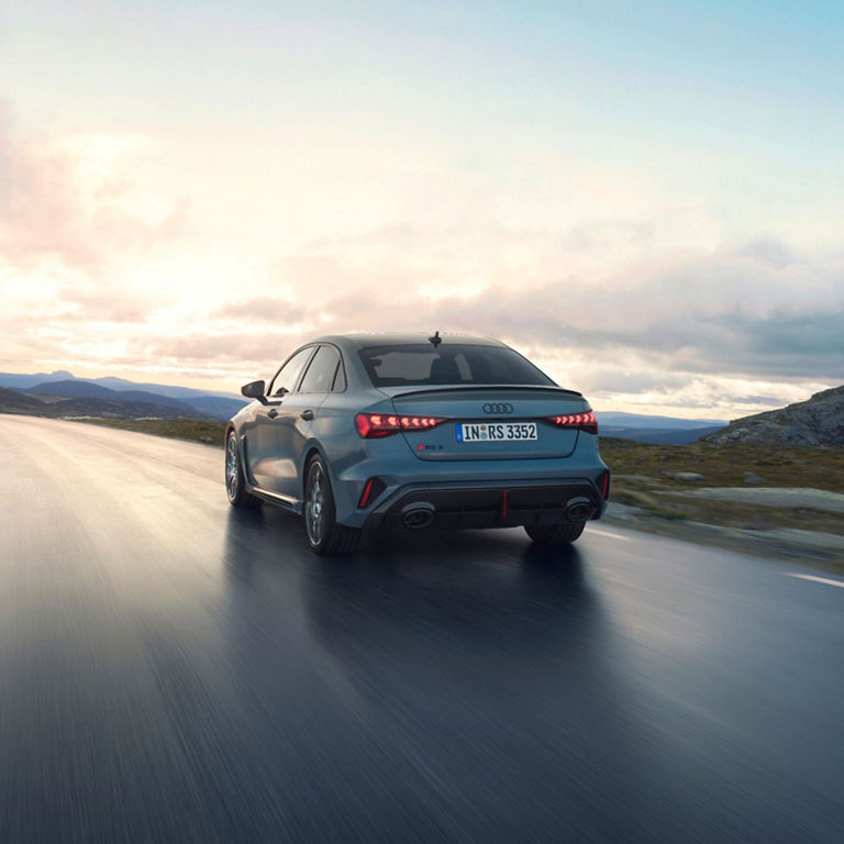 Blue Audi RS3 Saloon driving on a road with a cloudy sky background.