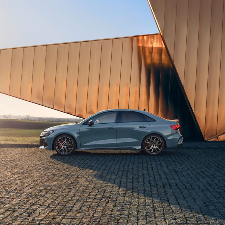 A blue Audi RS3 Saloon parked beside a large structure with a metallic copper finish at sunset.