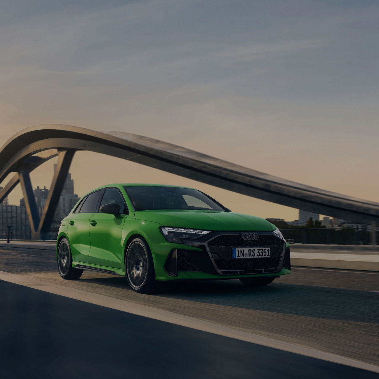 Green Audi S3 driving on bridge at dusk with city skyline in background.