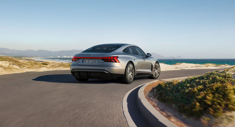 Silver Audi S e-tron GT car driving on coastal road with sea and mountains in the background.