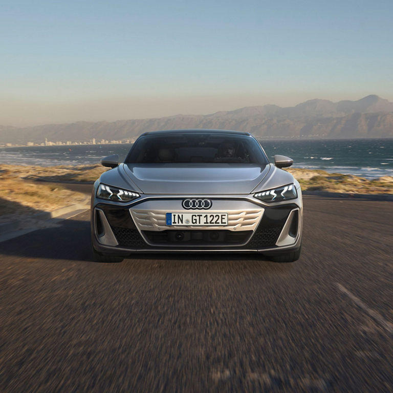 Silver Audi driving on coastal road with mountains and ocean in the background.