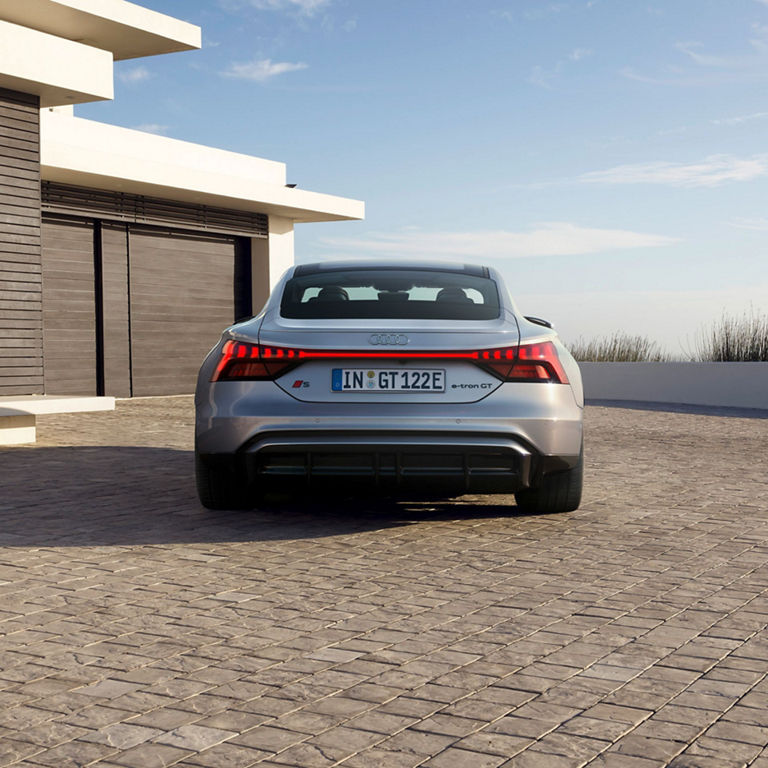 Silver Audi S-e-tron GT parked outside a modern house with a clear sky.