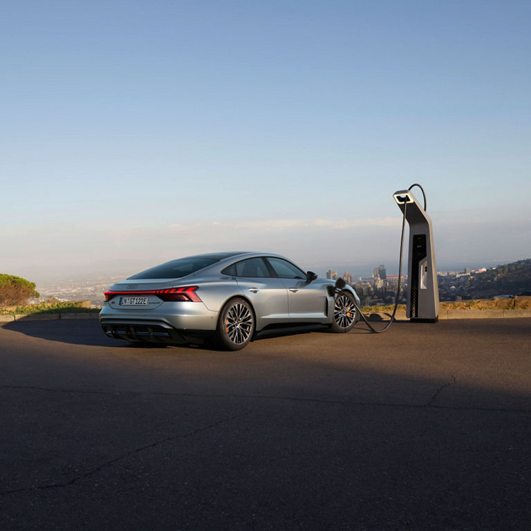 An electric Audi sedan charging at a public station overlooking a cityscape at dusk.