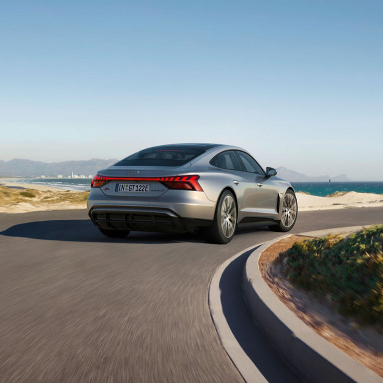 A silver Audi S-e-tron GT driving along a coastal road with mountains in the distance.