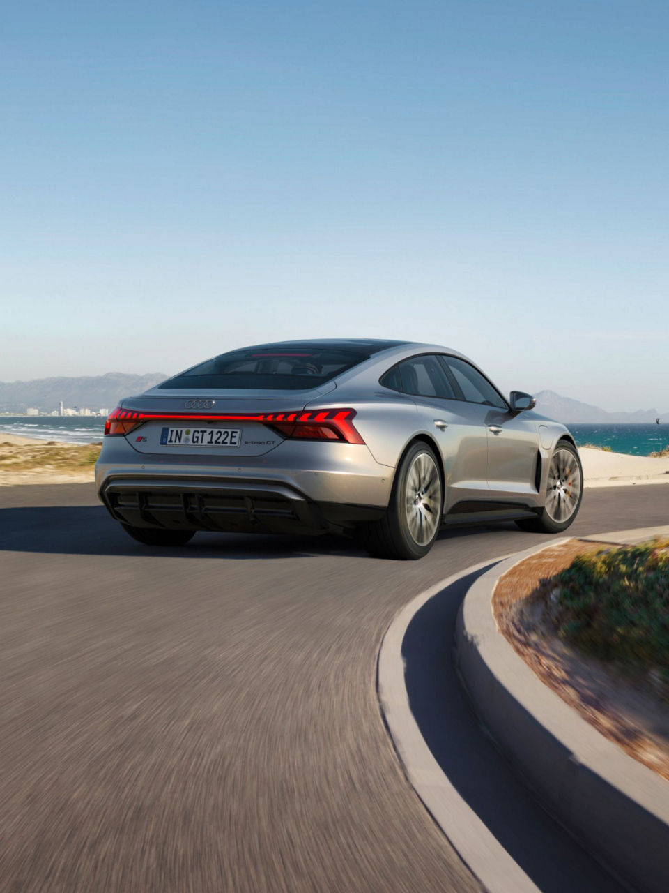 Silver Audi S e-tron GT car driving on coastal road with sea and mountains in the background.