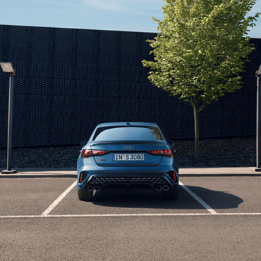 Blue Audi sedan parked outdoors near a tree and modern building facade.