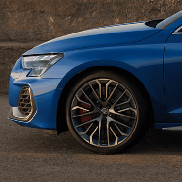 A close-up of a blue car's front left side, focusing on the wheel and headlight, against a concrete wall.