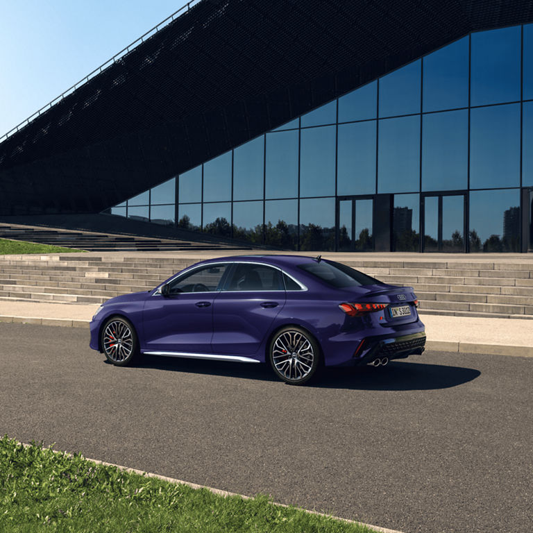 A purple Audi sedan parked beside a glass building under clear skies.