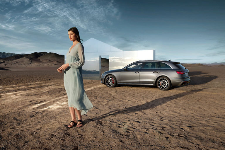 A person in a long dress stands by a modern car outside a glass house in a desert.