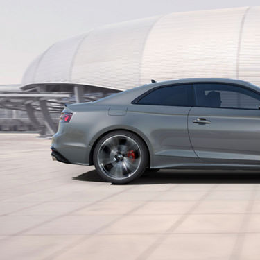 A grey coupe in motion on a smooth pavement with a blurred modern building in the background.