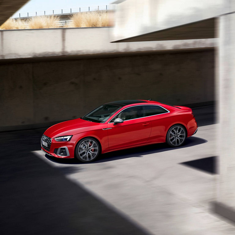 Red coupe parked along a sunlit concrete structure casting shadows.