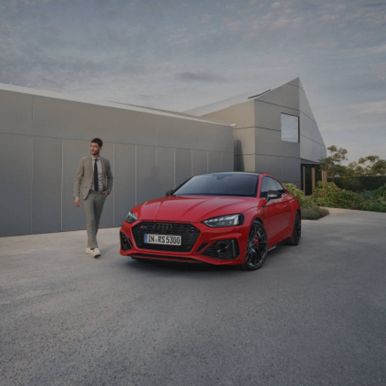 A person in a suit walking towards a red Audi car parked in front of a modern building.