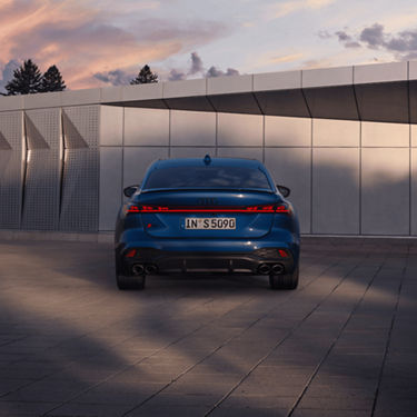 Blue Audi car parked with sunset and modern wall in background.