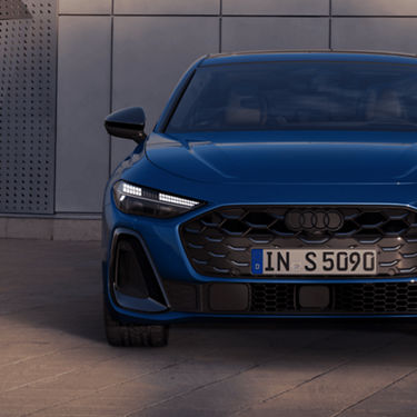 A blue Audi car parked in front of a grey wall with its headlights on.