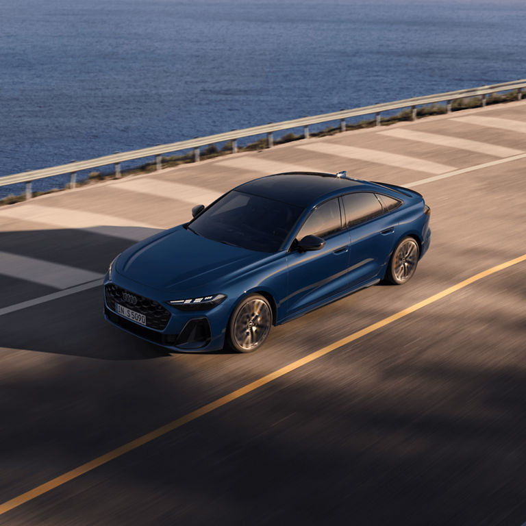 A blue sedan driving on a coastal road at sunset.